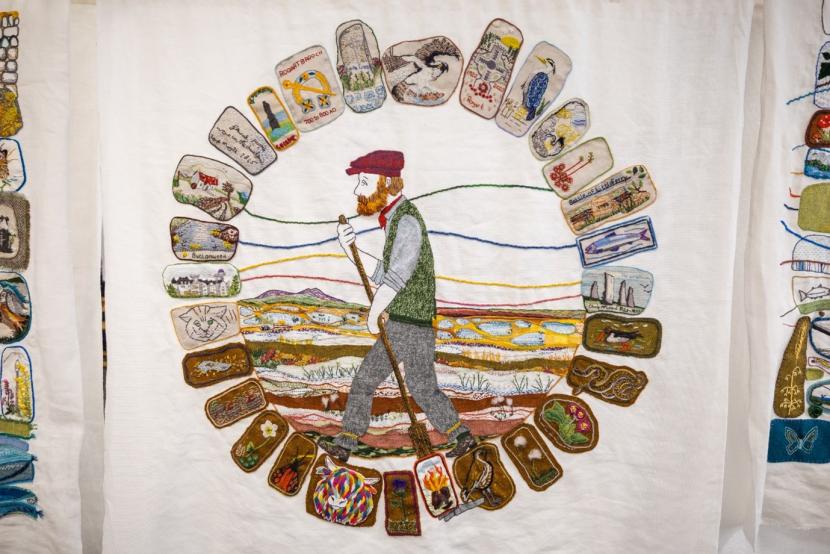 Tapestry hung vertically, showing the peat cutter and the landscape, surrounded by individual "stones" depicting a variety of local things.  