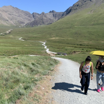 Two people walking on the Fairy Pools path; the Cuillin mountains in the background; one person with a yellow umbrella