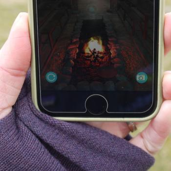 Uist Unearthed's app, on the Bornais site, showing a hand holding a phone, with the augmented reality image of a Viking Longhouse, and central fire.